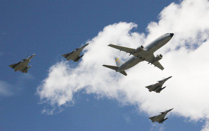 A Boeing 737, in the middle, is escorted by four IAI Kfir fighters, in an image seen from the bottom up with sky and clouds in the background. The aircraft belong to Colombia. Photo: Brazilian Air Force/EDA
