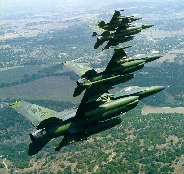 Foto mostra quatro caças F-16 com camuflagem verde sobrevoando área de campo.